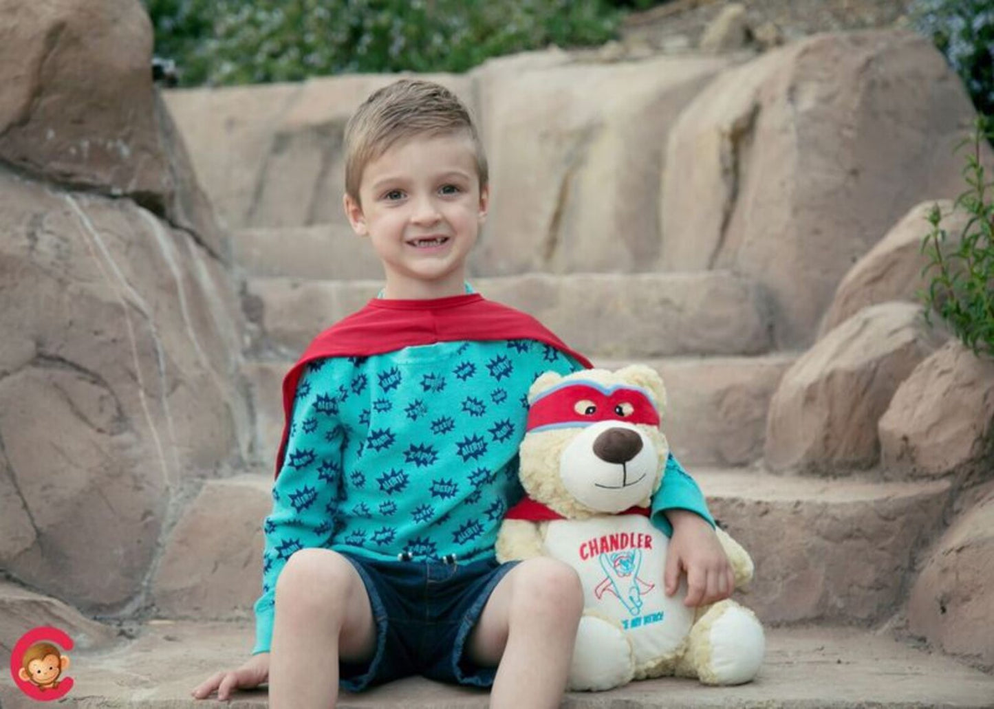 Child wearing a superhero costume with a red cape and holding a teddy bear on stone steps.