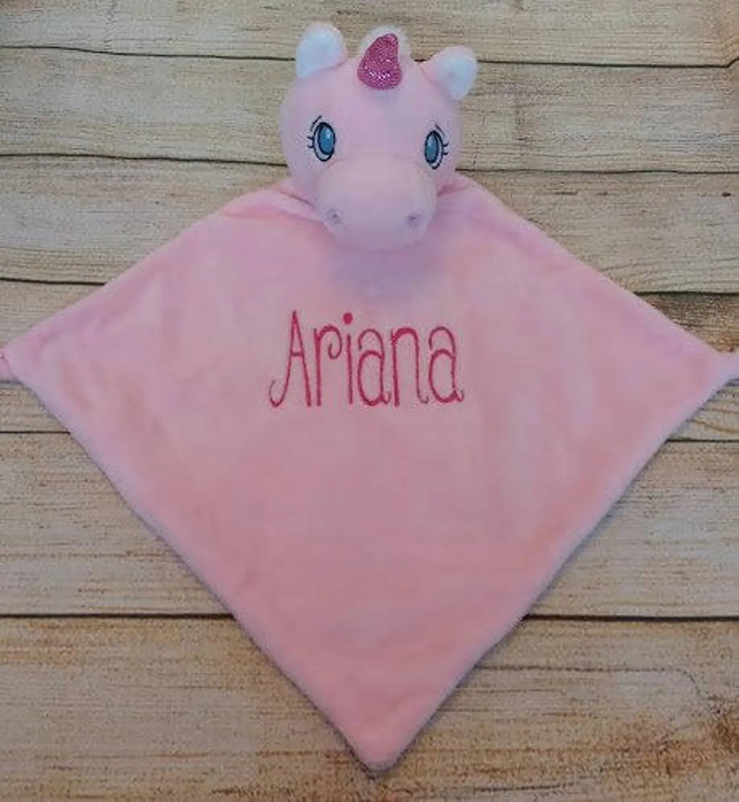 Pink plush toy with 'Ariana' embroidered on it, sitting on a wooden surface.