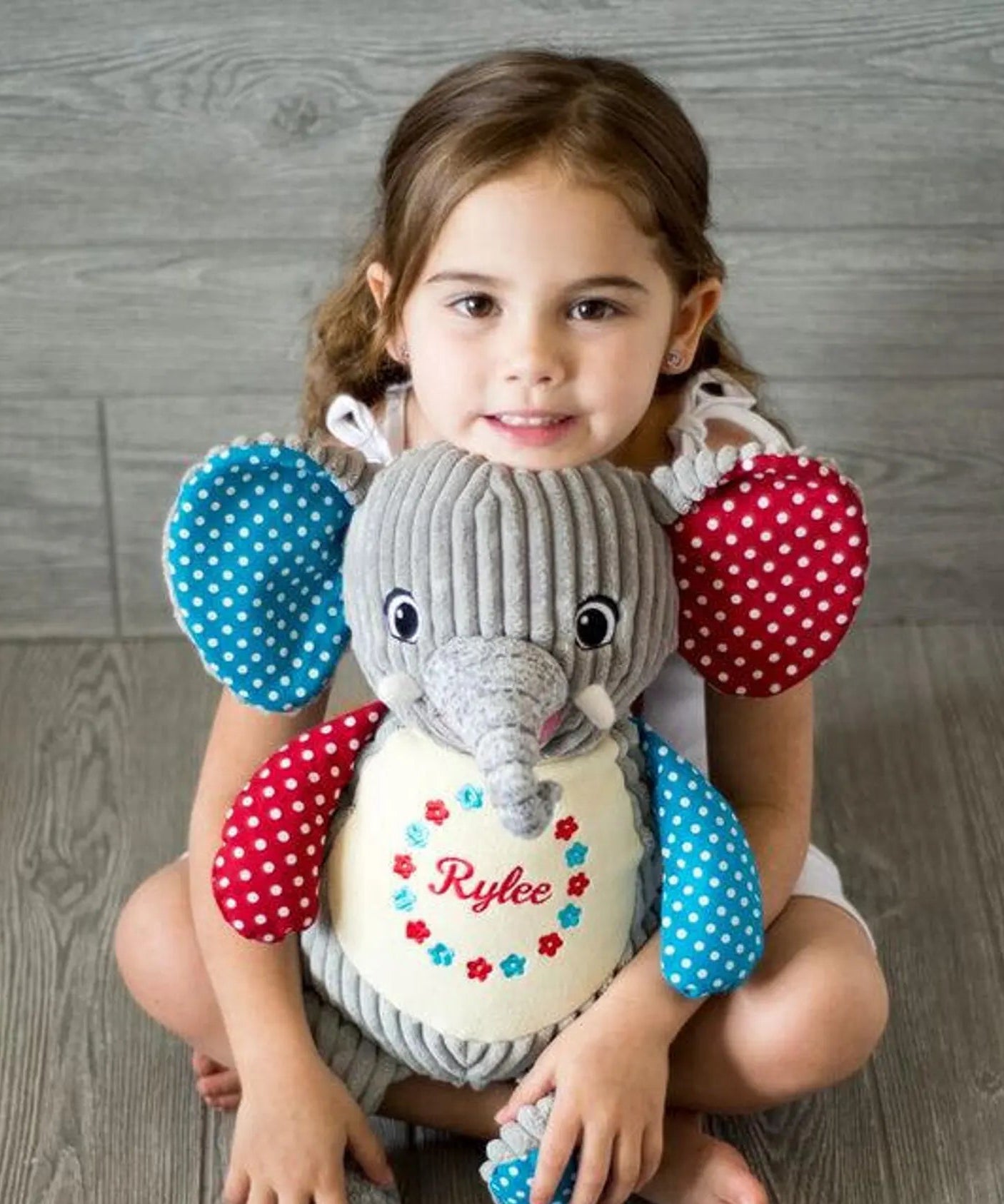 Young girl holding a personalized stuffed elephant toy with colorful sleeves on a wooden floor.