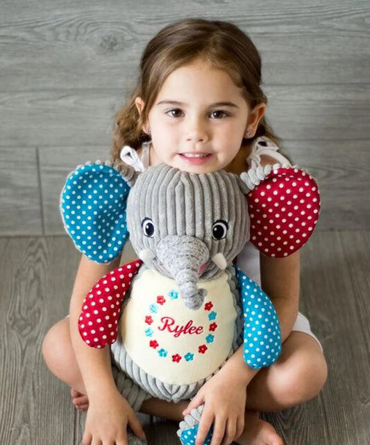 Young girl holding a personalized stuffed elephant toy with colorful sleeves on a wooden floor.