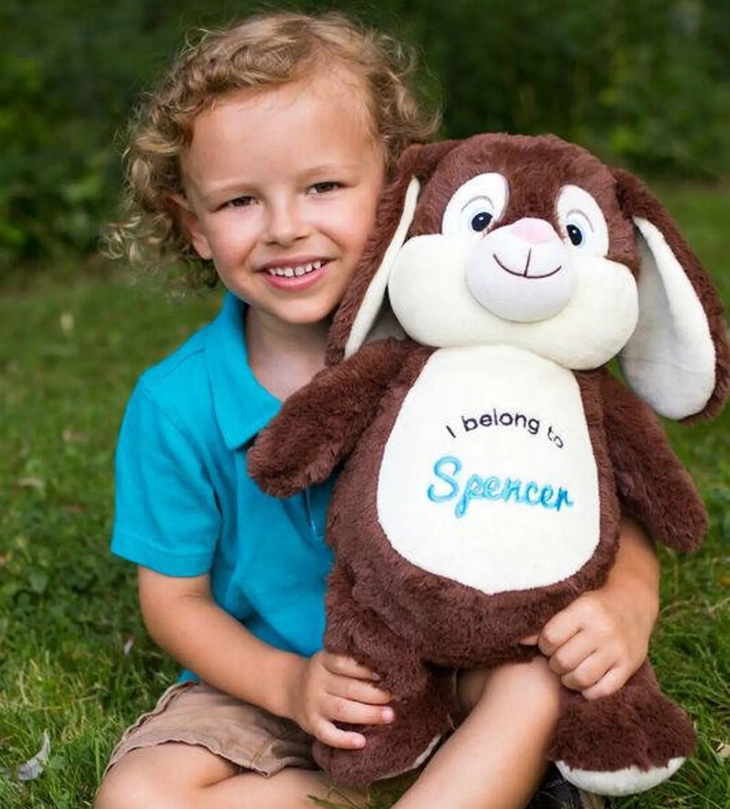 Child holding a plush toy with 'I belong to Spencer' text, sitting on grass.