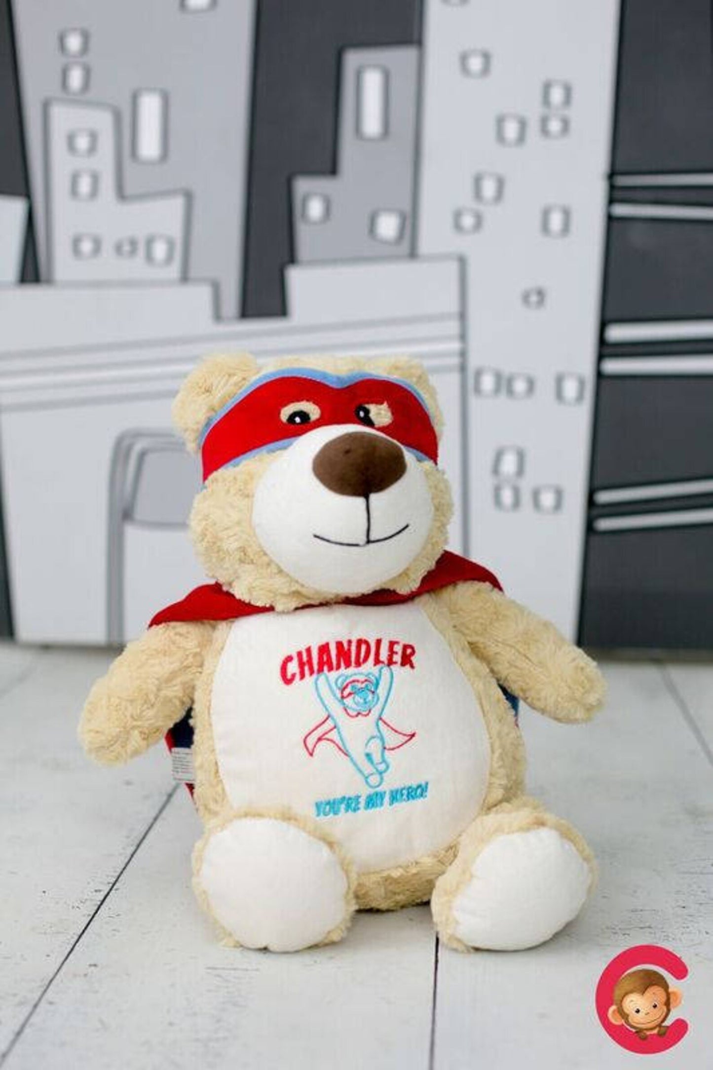 Teddy bear with a red cape and mask, against a cityscape background.