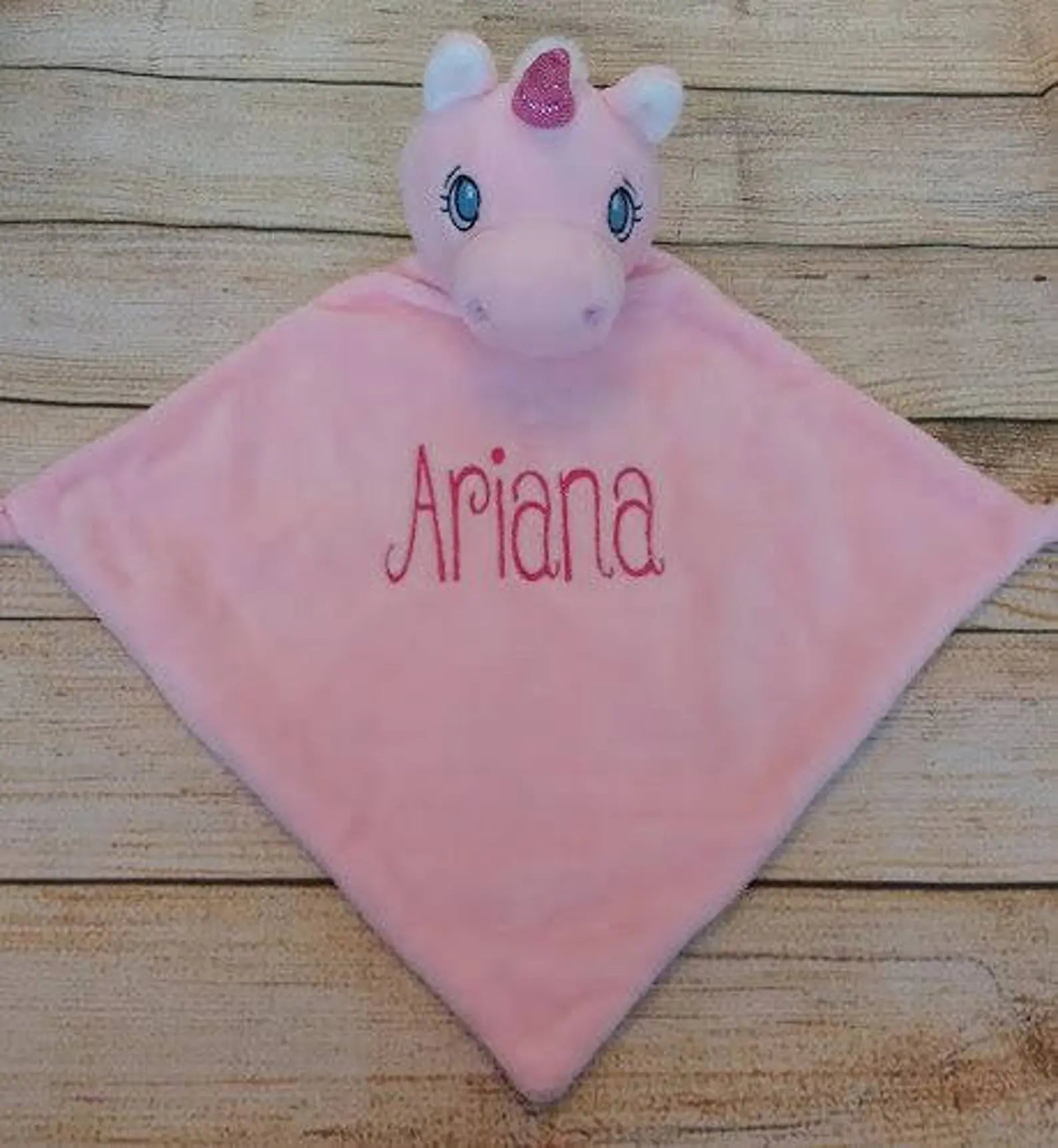 Pink plush toy with 'Ariana' embroidered on it, sitting on a wooden surface.