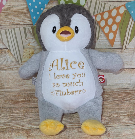 Plush penguin toy with 'Alice I love you so much Finbarr' text on a wooden surface with colorful flags in the background.