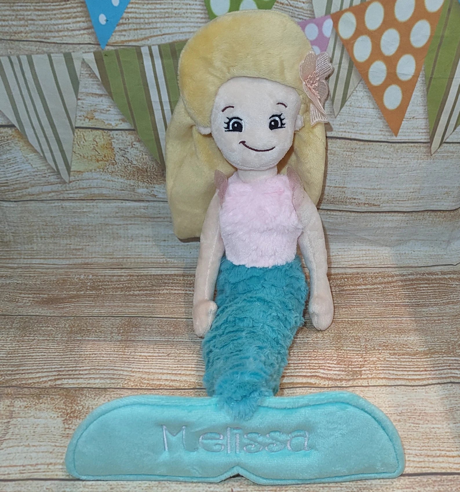 Plush mermaid toy with blonde hair and blue tail on a wooden surface, brand 'Melissa' visible.