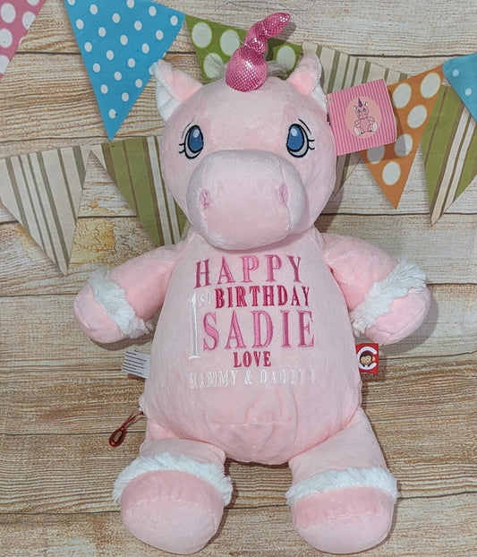 Pink unicorn plush toy with 'Happy Birthday Sadie' text on a wooden surface with colorful flags in the background.