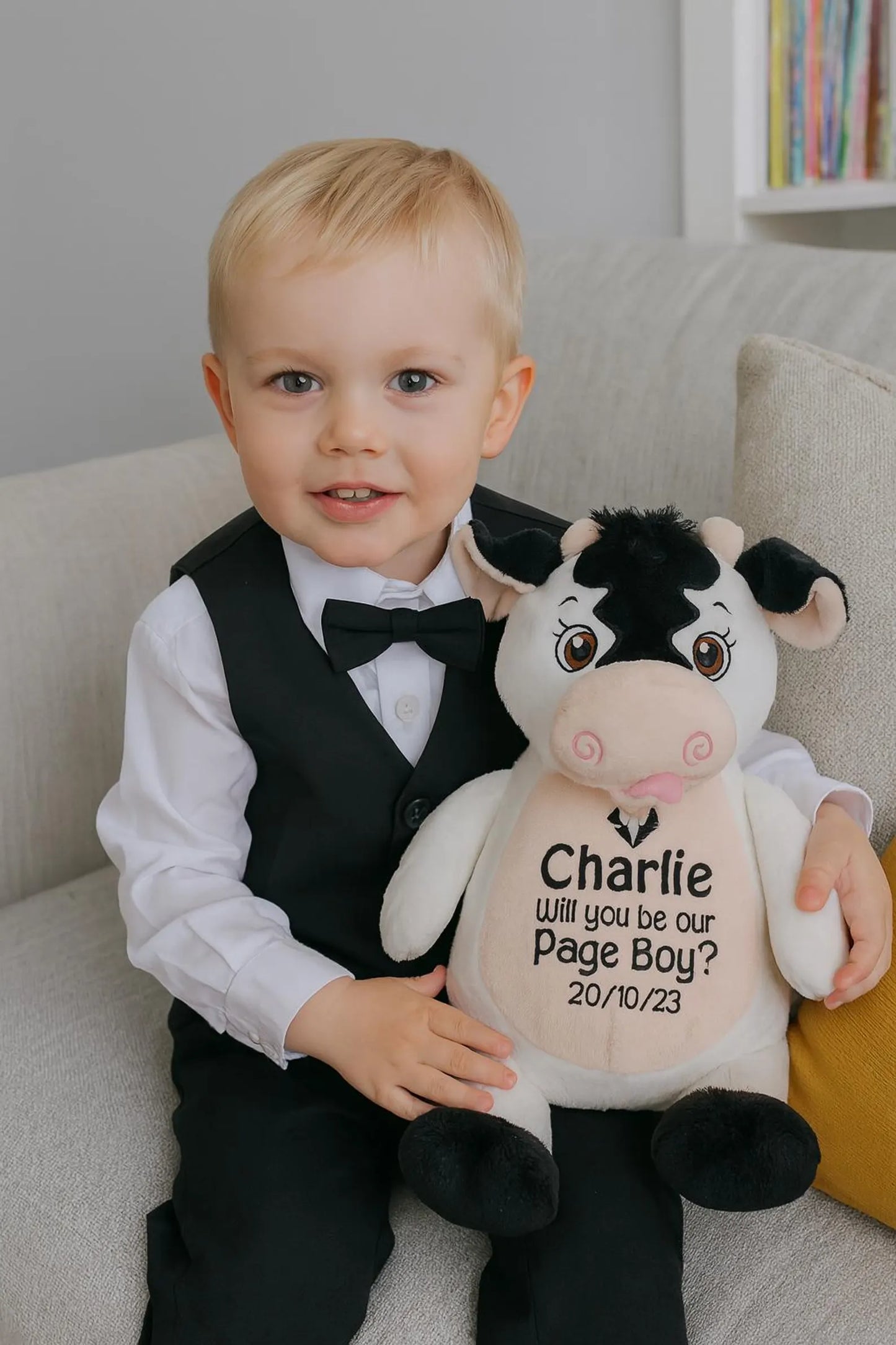 Young boy in formal attire holding a plush cow with personalized text, sitting on a couch.