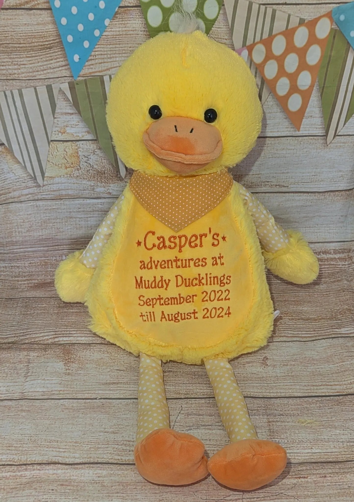 Yellow plush duck toy with text on a wooden surface and decorative flags in the background