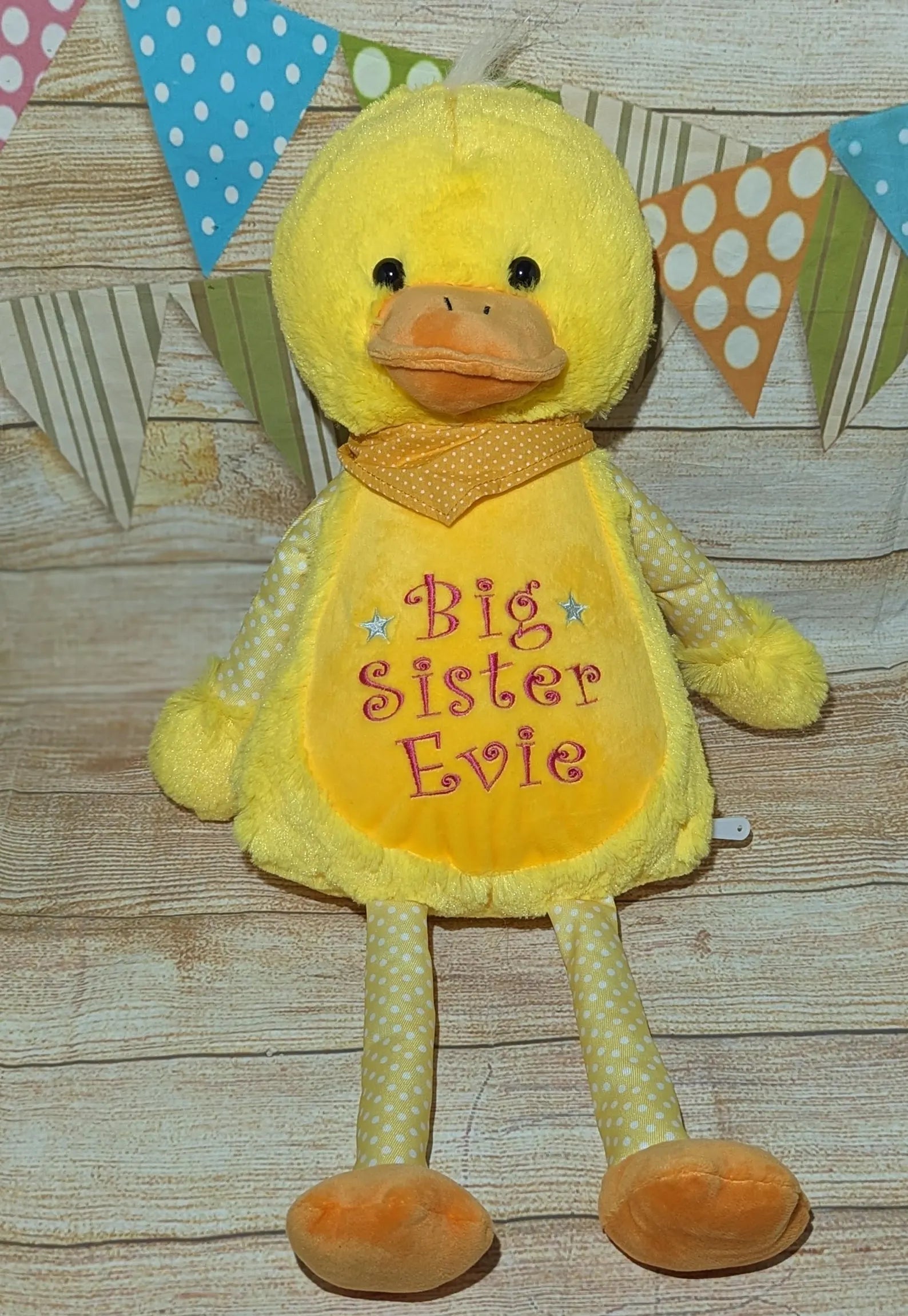 Yellow plush duck with 'Big Sister Evie' on a wooden surface with decorative flags in the background