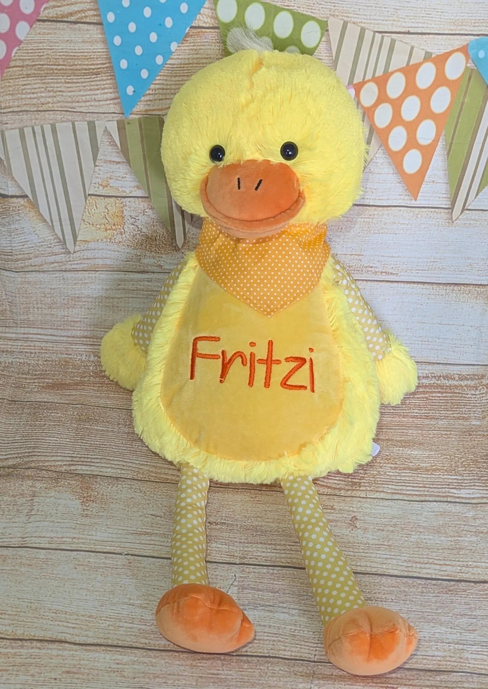 Yellow plush duck toy with 'Fritzi' embroidered on it, placed on a wooden surface with colorful flags in the background.