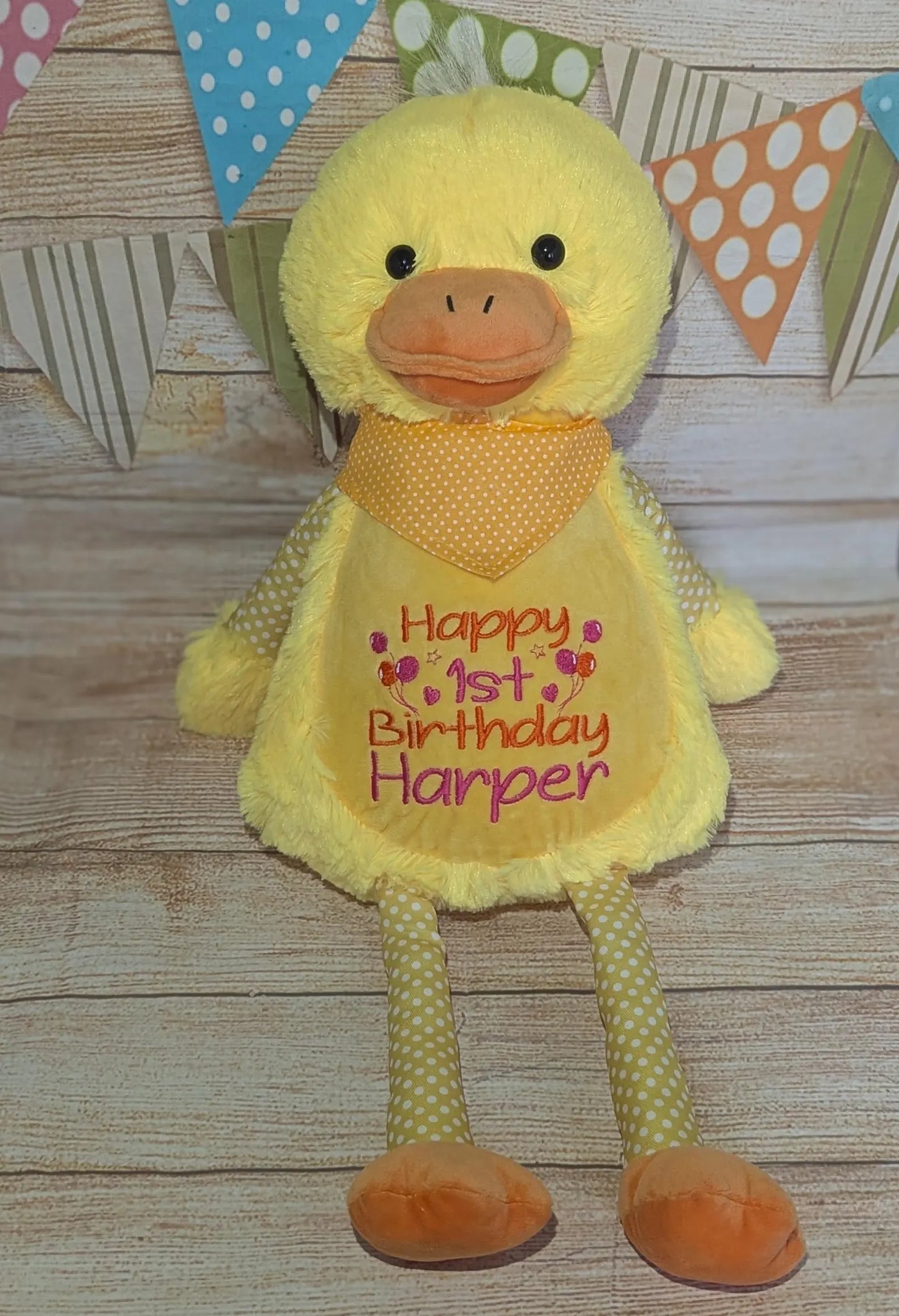 Yellow plush duck toy with a bib reading 'Happy 1st Birthday Harper' on a wooden surface with decorative flags.