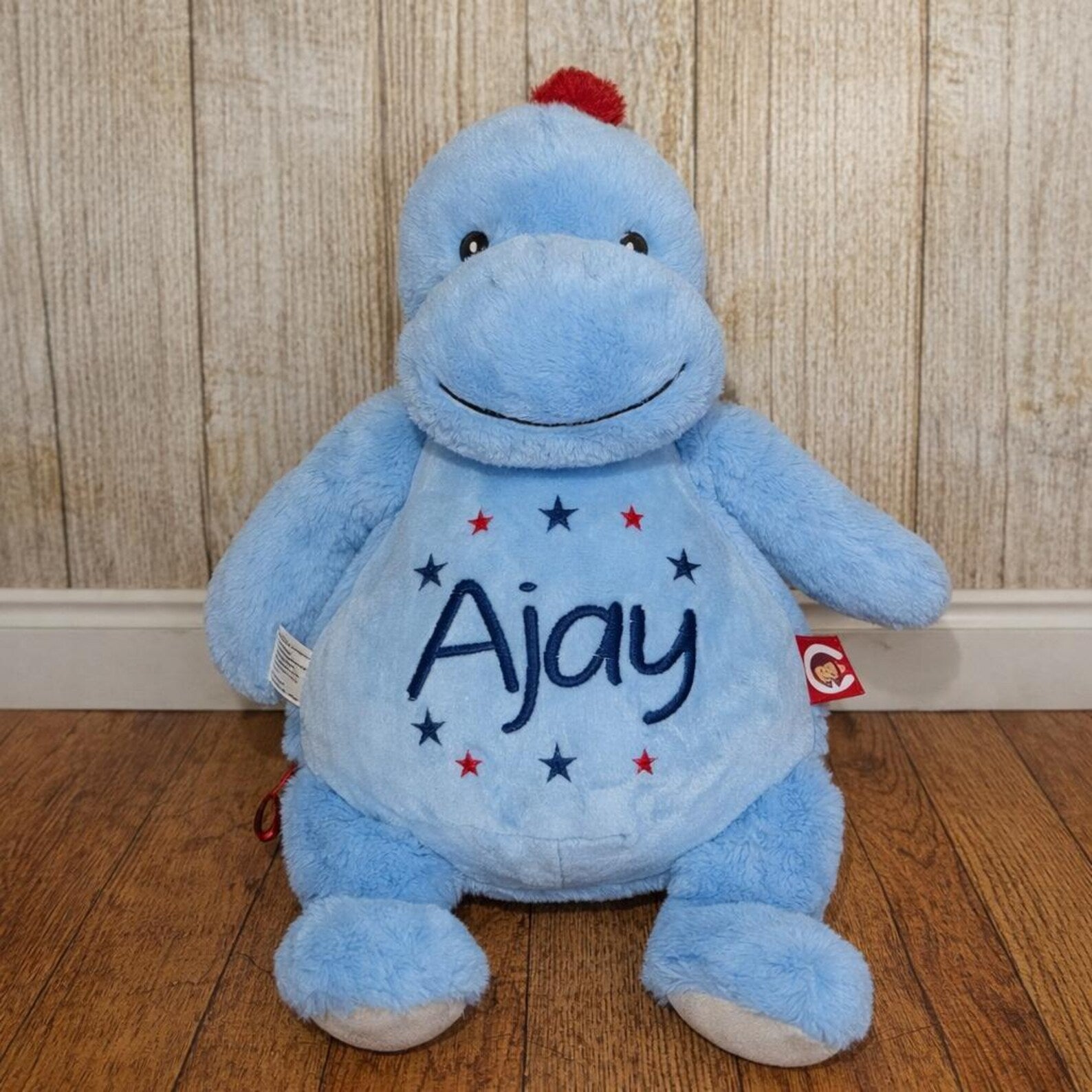Blue plush toy with 'Ajay' on a wooden floor and wooden wall background