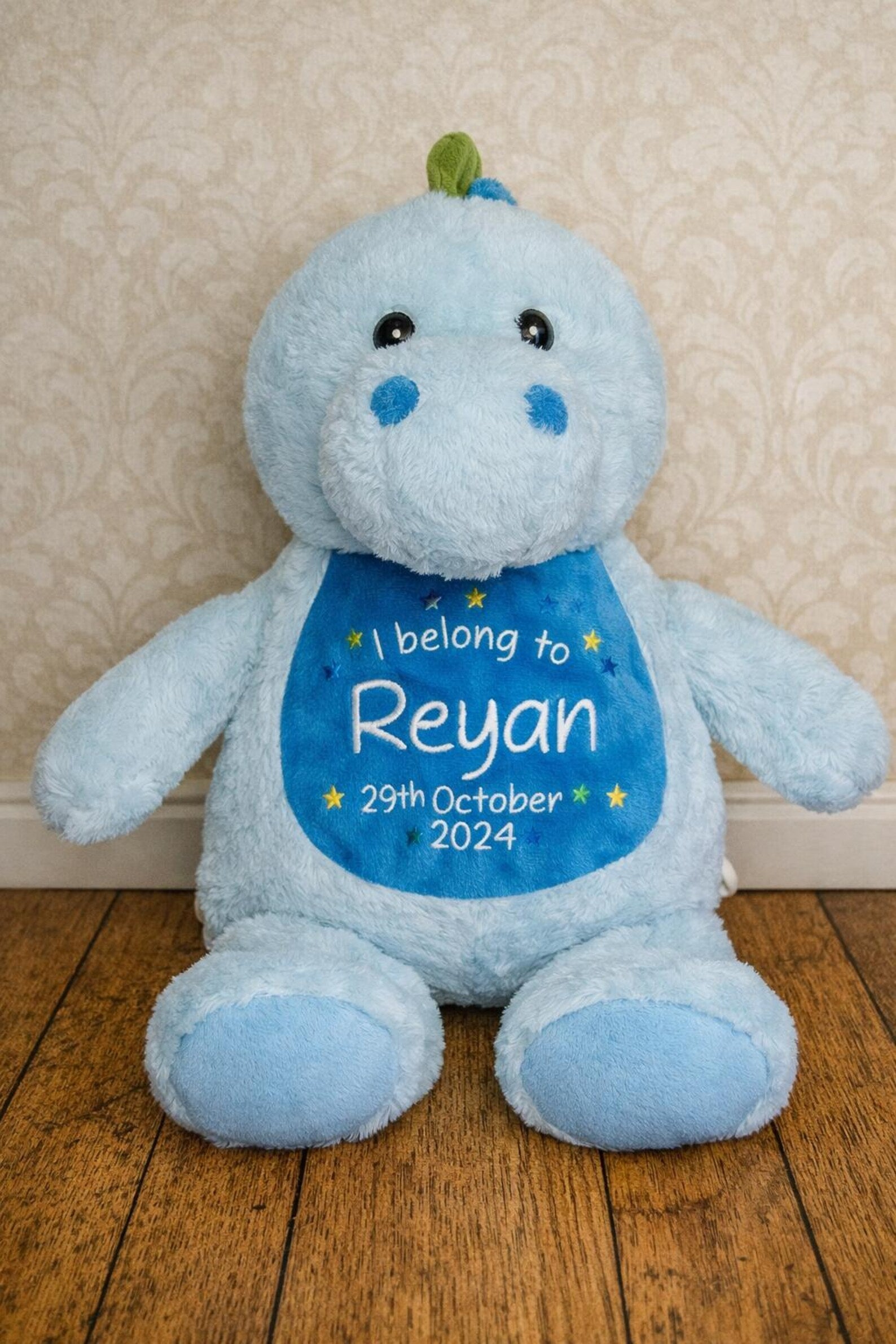 Blue plush toy with a name and date tag on a wooden floor against a beige wall.