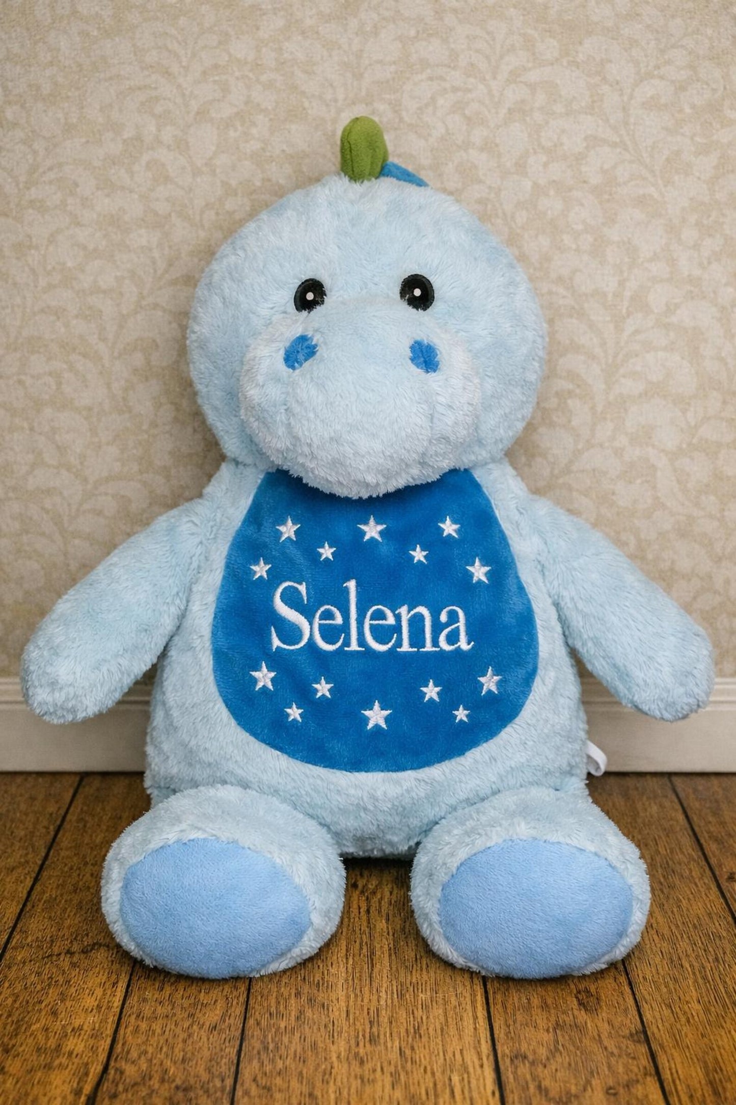 Blue plush toy with 'Selena' on a bib against a beige wall.