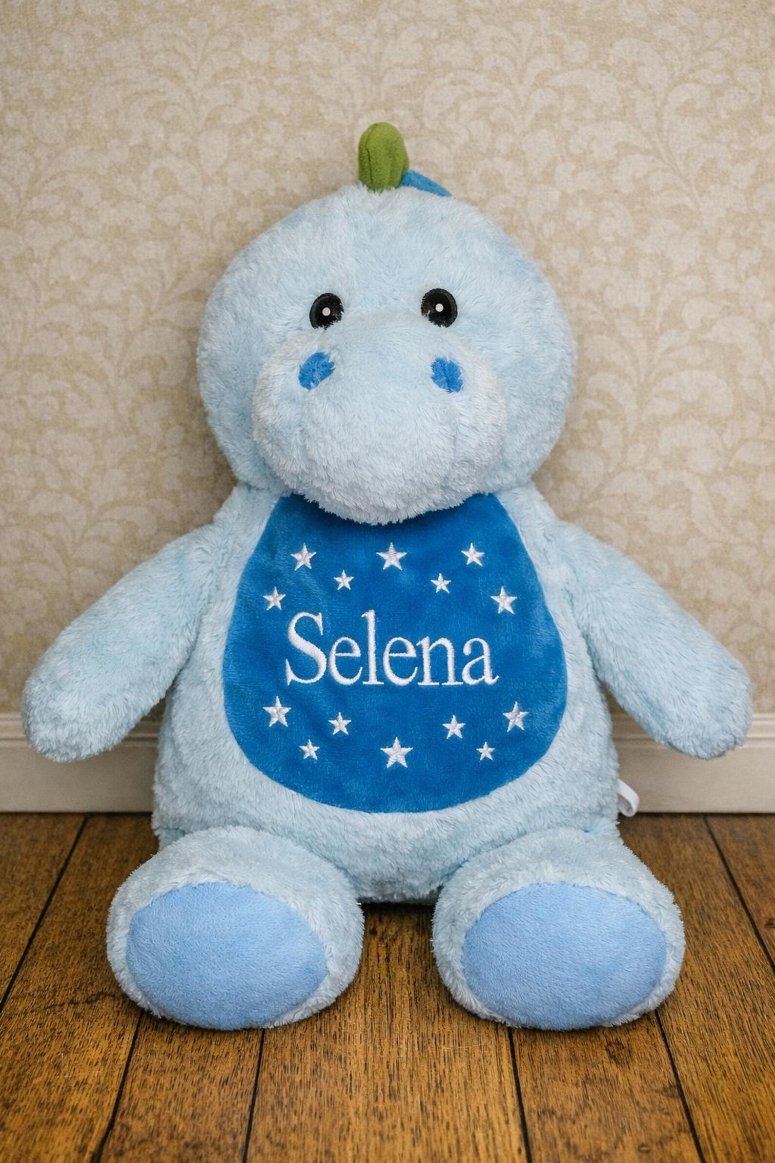Blue plush toy with 'Selena' on a bib against a beige wall.