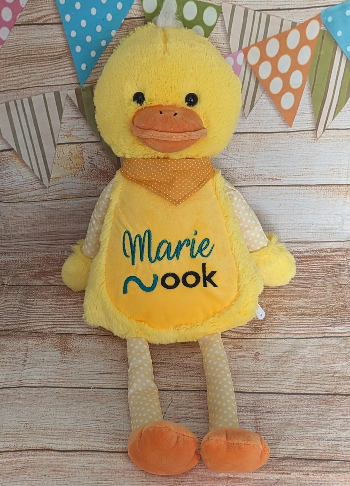Yellow plush duck toy with 'Marie Nook' on a wooden surface with colorful flags in the background