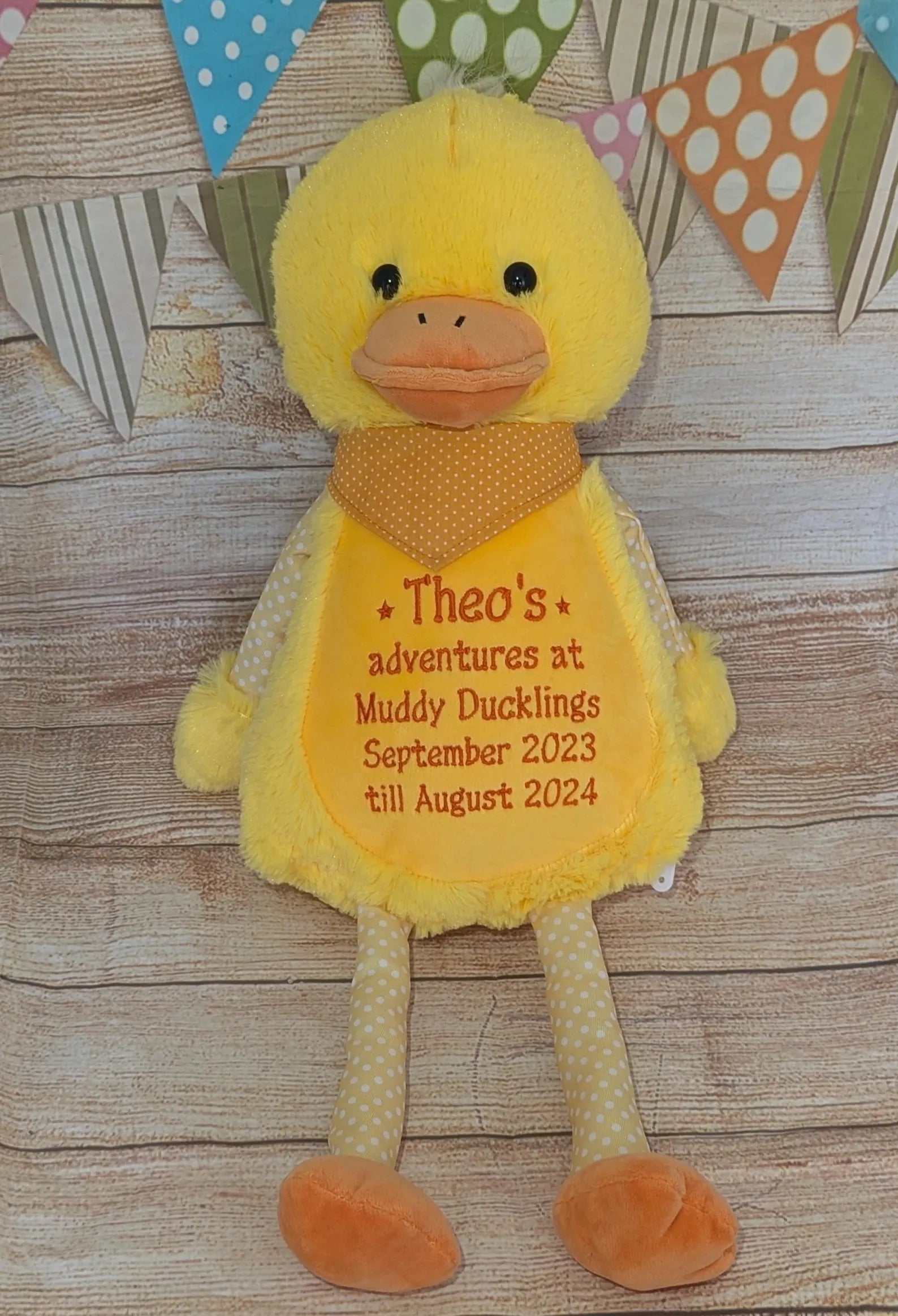 Yellow plush duck with text on a wooden surface with decorative flags in the background
