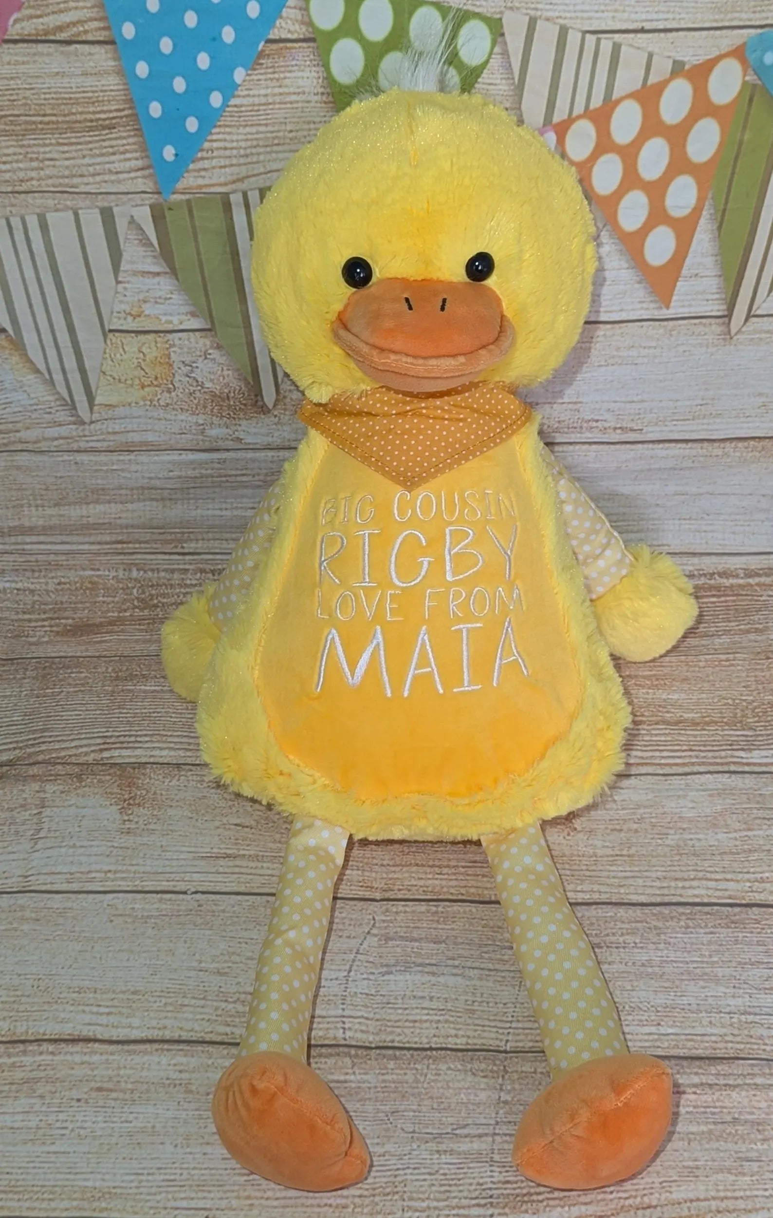 Yellow plush duck toy with text on a wooden surface and decorative flags in the background