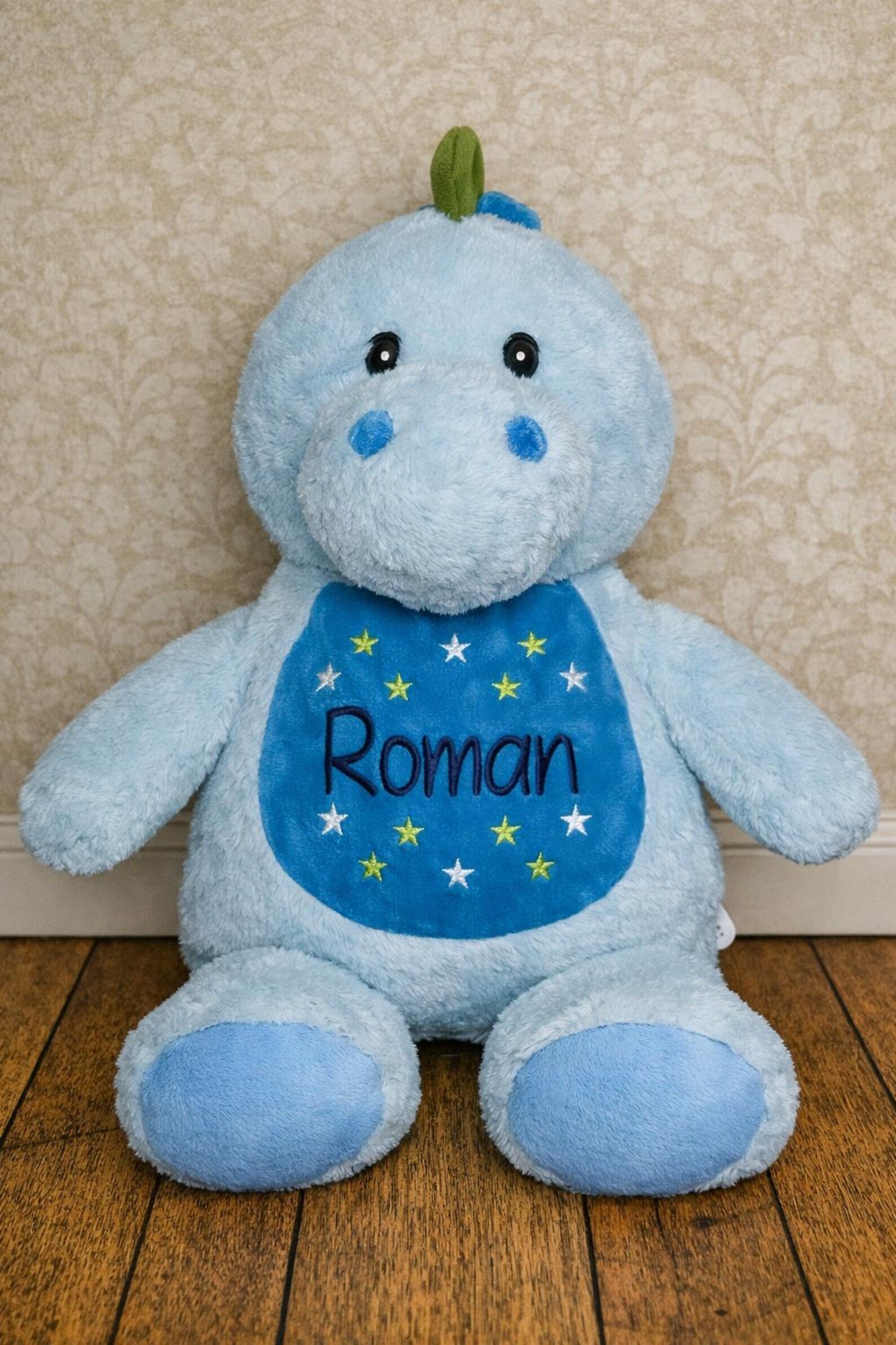 Blue plush toy with 'Roman' on a wooden floor against a beige wall.