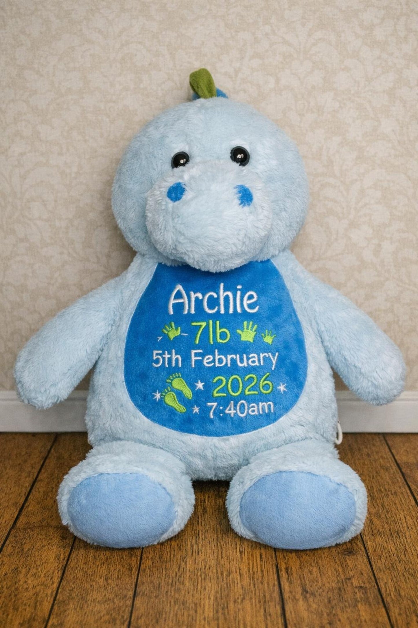 Blue teddy bear with personalized details on a wooden floor against a beige wall.