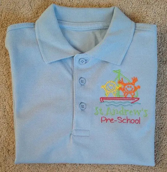 Folded light blue polo shirt with 'St Andrew's Pre-School' embroidery on a beige carpet.