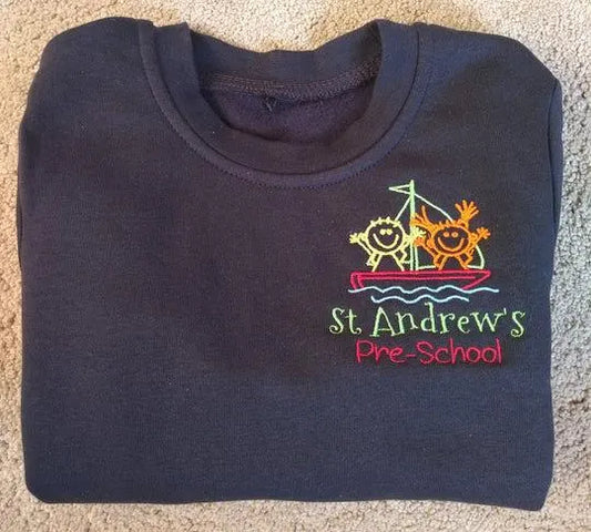 Folded navy blue shirt with 'St. Andrew's Pre-School' logo on a carpeted floor.